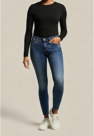 Femme portant un haut noir à manches longues, un jean skinny bleu et des baskets blanches, debout avec une main dans la poche.