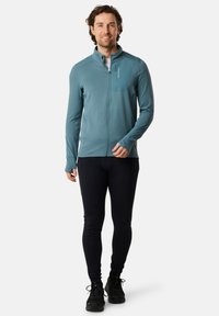 Miestens zils zipa žakets, kas izgatavots no teksturēta auduma, savienots ar melniem legingiem. Iespējas ietver kabatu uz krūtīm un trīs pirkstu caurumus piedurknēs.