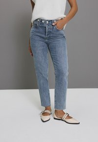 Jeans de culoare albastru deschis, cu talie înaltă și picior drept, având un sistem de închidere cu nasturi și buzunare frontale. Purtați cu pantofi joși crem cu accente negre și curele.