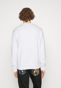 Felpa bianca con maniche lunghe e colletto rotondo, caratterizzata da una texture liscia e orlo a costine. Indossata con pantaloni neri con tasche decorate.