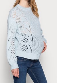 Ljusblå stickad tröja med ett öppet mönster, långa ärmar och avslappnad passform, kombinerad med mörkblå jeans.