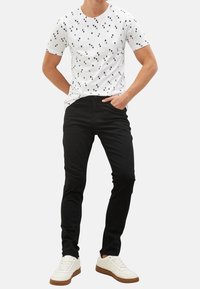 Bílá tričko s kulatým výstřihem a černým trojúhelníkovým vzorem, kombinované s úzkými černými kalhotami a bílými teniskami s hnědou podrážkou.