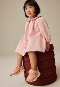 Lyserød stribet kjole med krave og knaplukning; barn iført beige og lyserøde sneakers med lilla detaljer, der sidder på en brun tekstureret skammel.