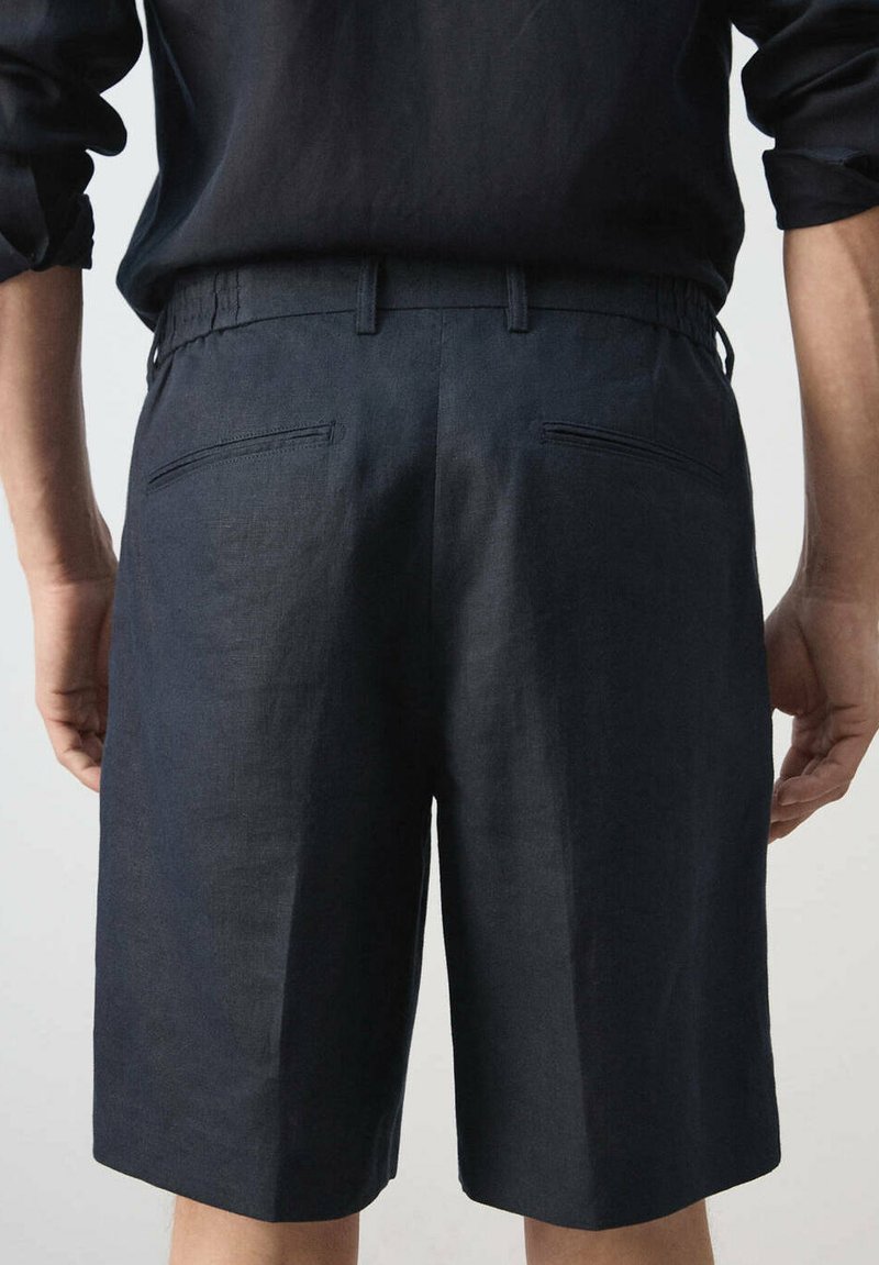 Personne portant un short bleu marine foncé sur mesure et une chemise sombre, vue de dos avec les mains détendues sur les côtés.