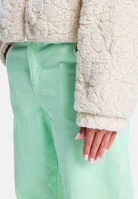 Kremowa kurtka o fakturze z liściastym wzorem, zestawiona z jasnozielonymi spodniami. Skupienie na fakturze materiału i subtelnym kontraście kolorów.