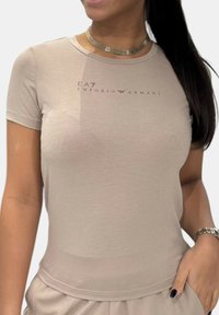 Camiseta ajustada beige de manga corta, con el logo de EA7 Emporio Armani impreso en un tono más oscuro en el pecho. Textura suave.