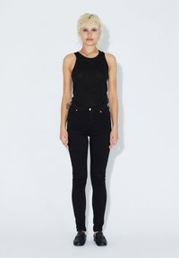 Černý bezrukávový top spárovaný s černými kalhotami s vysokým pasem. Látka se zdá hladká s minimem švů a subtilními detaily kovových prvků.