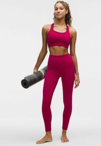 Fuksjowy stanik sportowy oraz legginsy z wysokim stanem o gładkiej fakturze. Modelka trzyma zwiniętą czarną matę do jogi. Stoi boso.