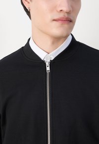Must musträkiga must zip-up jakk, millel on ümmargune kaelus, valmistatud sileda kanga. Omab hõbedast lukku ja nähtavat valget krae särk, mis on all.