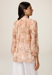 Blusa floreale in rosa chiaro, caratterizzata da maniche lunghe, un colletto a volant e delicati motivi floreali in tonalità di verde e rosa scuro.