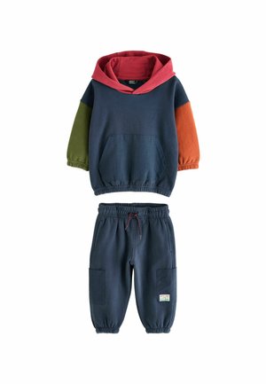 Kapucnis pulóver, multicolor ujjakkal (zöld, piros, narancs), tengerészkék joggingnadrággal párosítva. Elülső zsebével és elasztikus mandzsettáival rendelkezik.