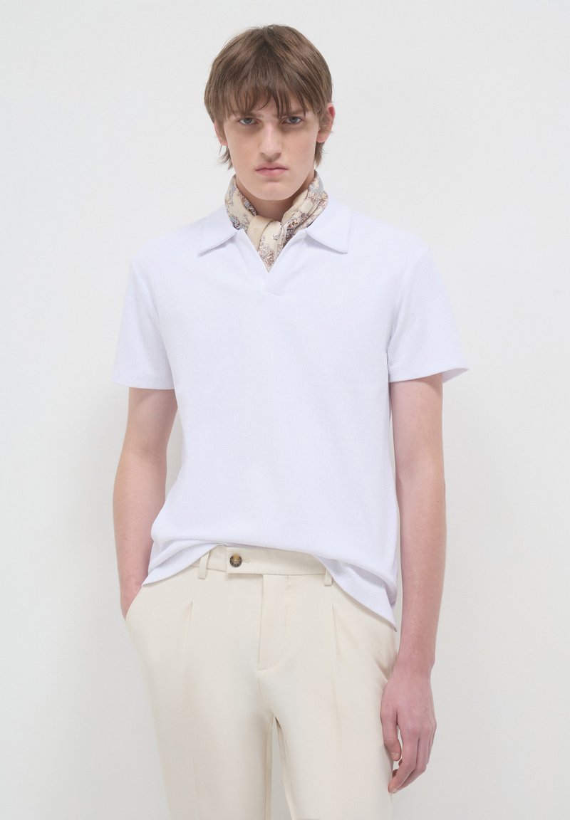 Polo shirt bianco a maniche corte con scollatura a V e tessuto a trama, abbinato a pantaloni beige chiaro su misura e una sciarpa a fantasia attorno al collo.