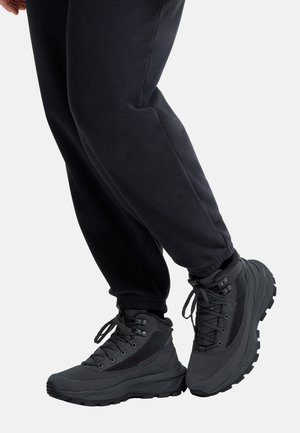 Persoon draagt zwarte stevige wandelschoenen en een zwarte broek, staat met één voet licht opgetild tegen een effen achtergrond.