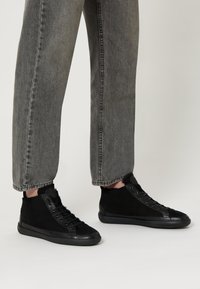 Sneaker alte nere in suede con suole in gomma, caratterizzate da un design con lacci e dettagli minimalisti neri. Indossate con jeans grigi scoloriti.