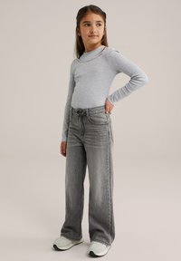 Graues langarmiges Oberteil mit subtilen Verzierungen, kombiniert mit weiten, verwaschenen grauen Jeans. Das Model trägt helle Sneakers und hat die Hände auf die Hüften gestützt.