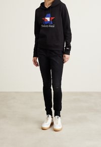 Černá mikina s barevným logem hvězdy, černé džíny s opotřebovaným vzhledem a bílé tenisky s gumovými podrážkami. Neformální outfit s moderními designovými prvky.