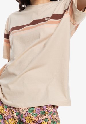 Beżowy T-shirt z brązowymi i różowymi ukośnymi paskami oraz logo, noszony z kolorowymi spodniami w kwiatowy wzór.