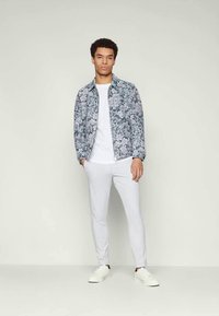 Ung mand iført en grå blomsterjakke, hvid t-shirt, lysegrå bukser og hvide sneakers, der står foran en ensartet lys baggrund.