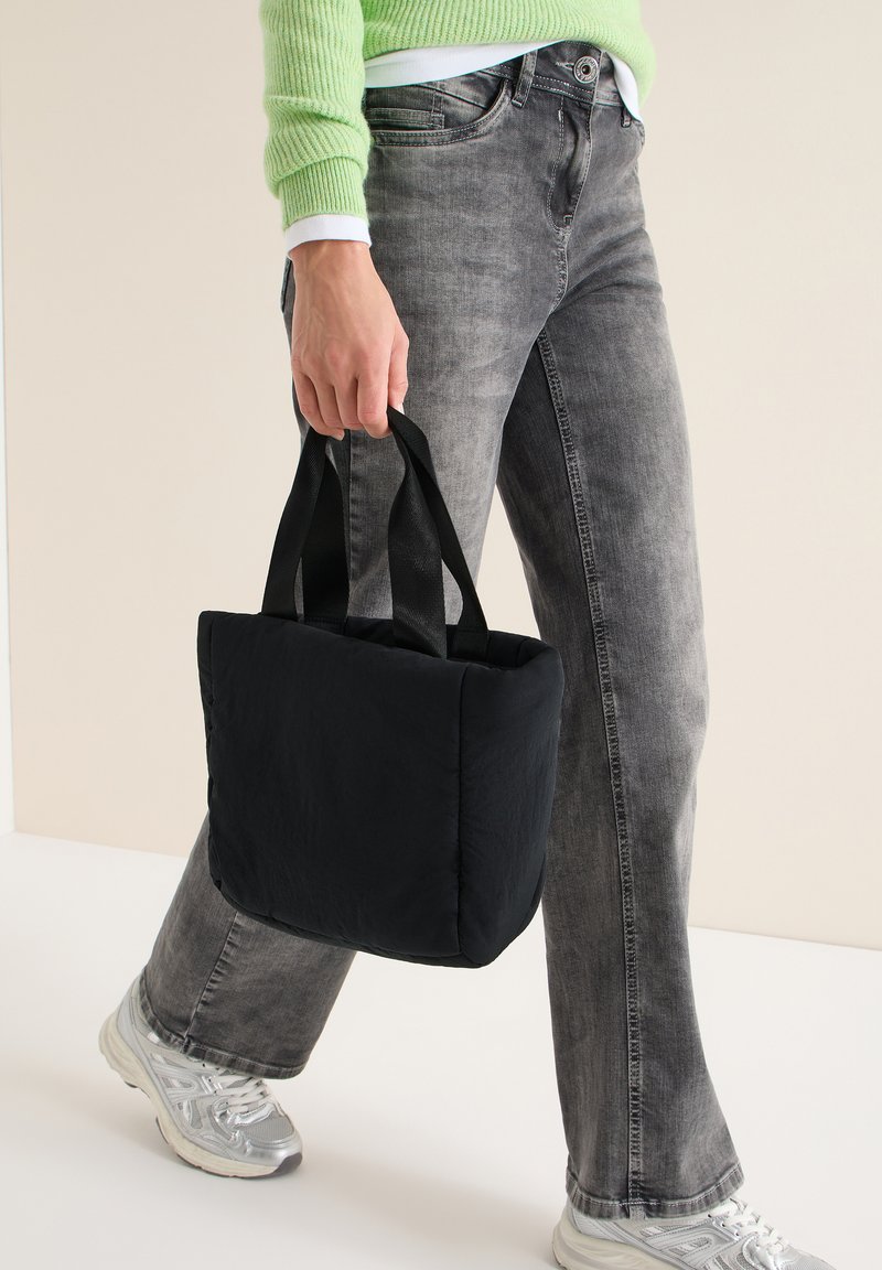 Sac fourre-tout matelassé noir avec deux poignées noires, porté par une personne. Le sac présente une forme rectangulaire et une texture lisse, légèrement brillante.