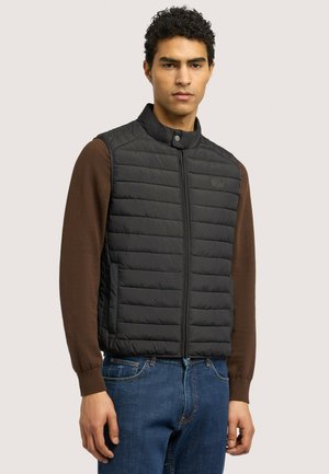 Joven con un chaleco negro acolchado sin mangas sobre una camisa marrón de manga larga y vaqueros azules, de pie frente a un fondo liso.