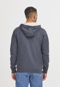 Sweatshirt à capuche bleu marine avec une texture douce, comportant une poche kangourou, des poignets et un ourlet côtelés. Porté par-dessus une couche plus claire.