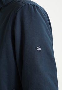 Námořnická modrá bunda s hladkou texturou, která má malé černé logo na levém rukávu a strukturovaný design límce.