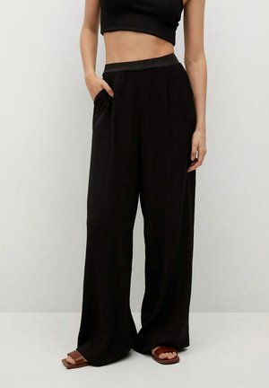 Femme portant un pantalon noir à jambes larges avec une taille élastique, main dans la poche, associé à des sandales marron à bout carré et un haut noir court.