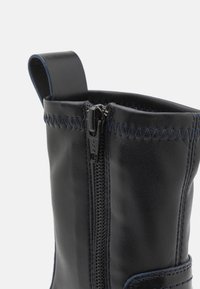 Stivale in pelle nera con chiusura a zip, cuciture blu a contrasto e un tab di estrazione a forma di anello nella parte superiore. Presenta una finitura liscia e texture.