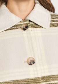 Rutigt röd med en kräm- och ljusgrön mönstring, mjuk texturerad krage och stora, cirkulära knappar med ett virvlande mönster.