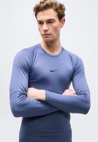 Νεαρός με σγουρά μαλλιά που φοράει στενή μπλε μπλούζα Nike με μακριά μανίκια, με σταυρωμένα τα χέρια, κοιτάζει προς τα κάτω και προς το πλάι σε απλό φόντο.