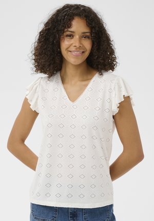 Kobieta o kręconych włosach, ubrana w biały wzorzysty top z dekoltem w serek i falbankami na rękawach oraz niebieskie dżinsy, uśmiechająca się z rękami za plecami.