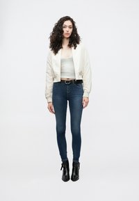 Chaqueta bomber blanca, top corto gris claro, vaqueros ajustados azul oscuro, cinturón negro y botines hasta el tobillo. Tejido liso y diseño ajustado.