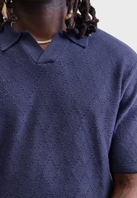Cămașă polo cu mânecă scurtă de culoare bleumarin, cu decolteu în V, cu un model texturat în formă de diamant și material tricotat. Dispune de o guler clasic și un tiv îngrijit.