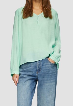 Vrouw draagt lichtgroene blouse met lange mouwen en knoopjes aan de V-hals, blauwe spijkerbroek, hand in de pocket, tegen een effen achtergrond.