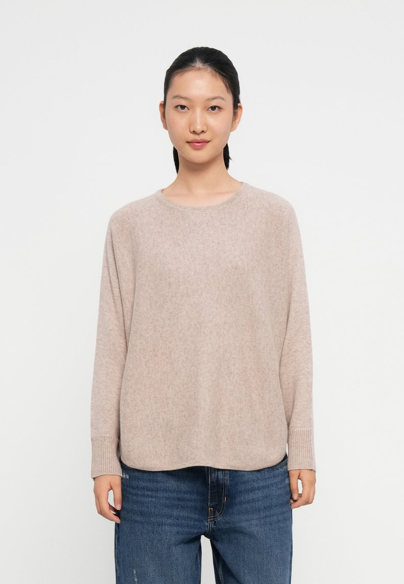 Light beige knitted sweater with long sleeves, a round neckline, and ribbed cuffs, paired with blue denim jeans. Soft texture and relaxed fit.