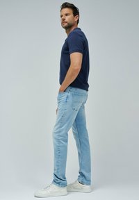 Polo marino de manga corta, jeans azul claros y zapatillas blancas. El modelo está de lado con una mano en el bolsillo contra un fondo liso.