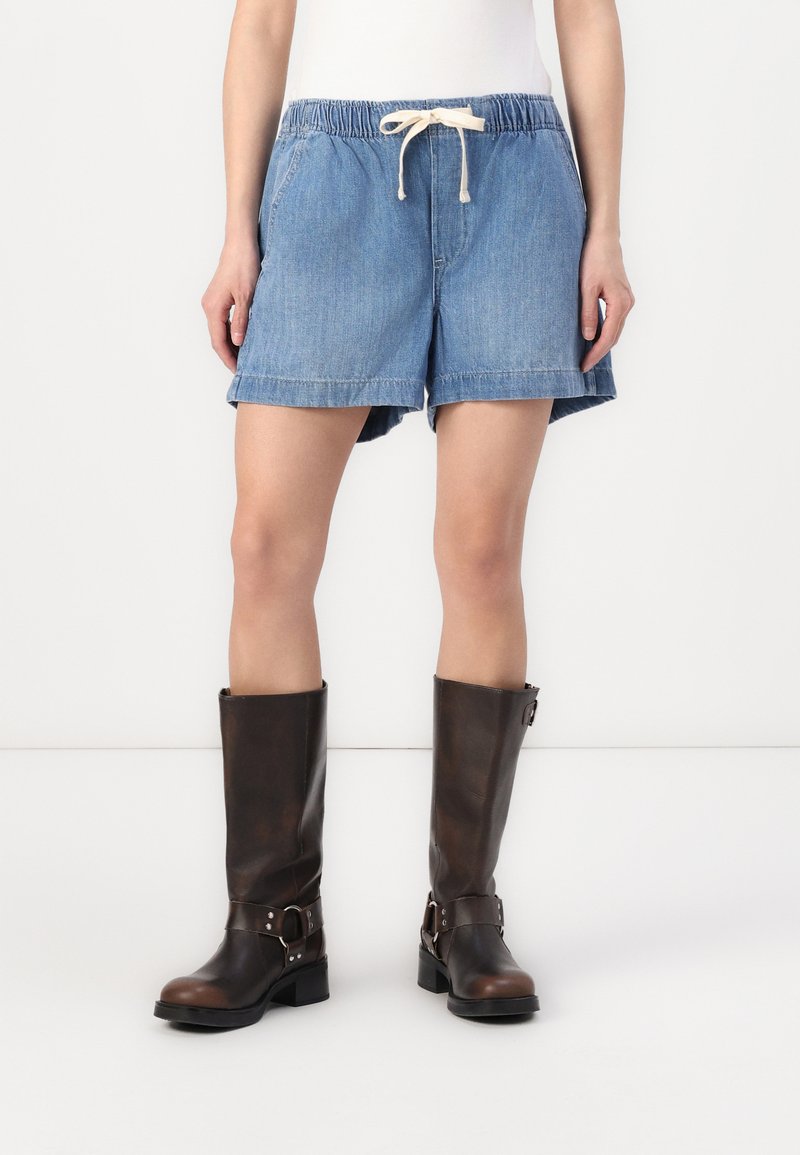 Jeans šortai su elastinga juosmeniu ir raišteliu, šviesiai mėlynos spalvos, su šoninėmis kišenėmis ir laisvu kirpimu, derinami su rudiems kelių ilgių batais.