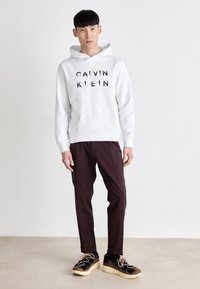 Bela kapuca z napisom "CALVIN KLEIN" v črni barvi, v kombinaciji s temno burgundskimi hlačami in črnimi teniskami s svetlo rjavimi akcenti in vezalkami.