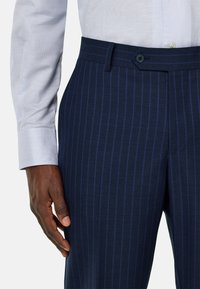 Pantalons rayés bleu marine avec une texture lisse, dotés d'une fermeture à un bouton et d'une coupe fuselée. Une chemise en tissu bleu clair est partiellement visible.