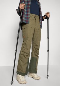 Persoon in olijfgroene wandelschoenen en beige wandelschoenen houdt twee zwarte trekkingstokken vast tegen een effen achtergrond.