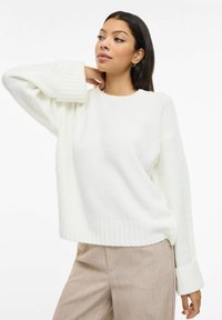 Biały dziergany sweter z żebrowanymi mankietami i dołem, o luźnym fasonie i miękkiej fakturze, w połączeniu z jasnobeżowymi spodniami.