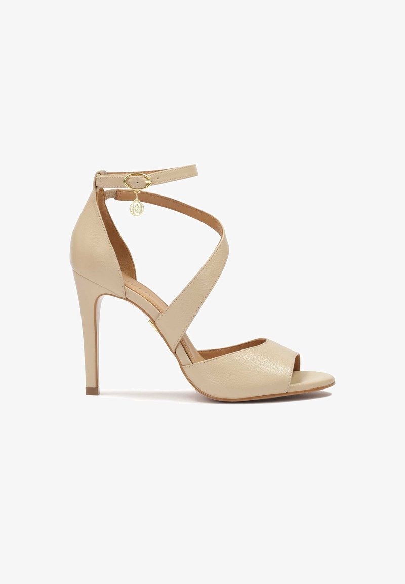 Sandales à talons hauts en cuir beige avec bout ouvert, brides croisées à la cheville et charm doré en accent. Texture lisse et design simple.