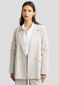 Vaalean beige kaksirivinen bleiseri, joka on valmistettu pellavasta ja siinä on rento fit. Bleiserissä on suuret napit ja etutaskut, ja se on yhdistetty valkoiseen paitaan.