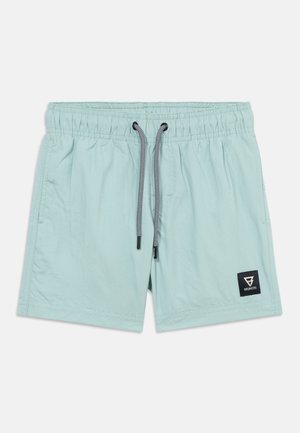 Bañador de hombre azul claro con cintura elástica, cordón negro y blanco, bolsillos laterales y parche con logo negro en la pierna izquierda.