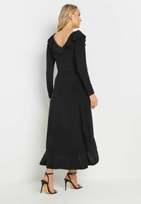Długa czarna sukienka z dekoltem w kształcie litery V, falbaniastym kołnierzem i długimi rękawami. Wykonana z gładkiego materiału; ma rozkloszowany dolny brzeg. Noszona z czarnymi sandałami na obcasie.