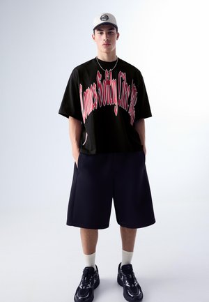 Young man wearing a white cap, oversized black T-shirt with red text, loose navy shorts, white socks, and black sneakers against a plain background.