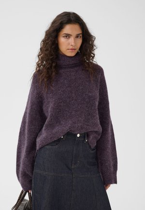 Femme aux longs cheveux bouclés portant un pull à col roulé violet et une jupe en jean foncé, tenant un sac à main devant un fond uni.