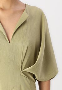 Rochie de un verde măsliniu cu decolteu adânc în V, mâneci scurte și o țesătură texturată. Prezintă un detaliu plisat la talie pentru a oferi formă.