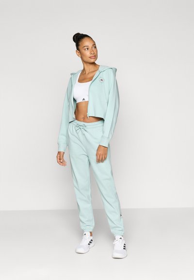 Ανοιχτό πράσινο μίντ cropped φούτερ με φερμουάρ και λογότυπο, σετ με ταιριαστά τζόγκερ με ελαστική μέση και μανσέτες. Λευκά αθλητικά παπούτσια.