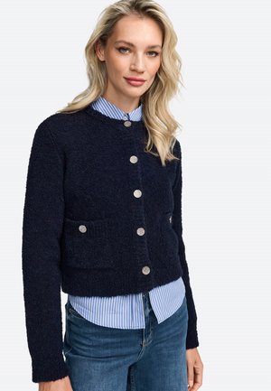 Cardigan corto blu navy in tessuto strutturato, con bottoni argentati e due tasche frontali, indossato sopra una camicia a righe azzurre.